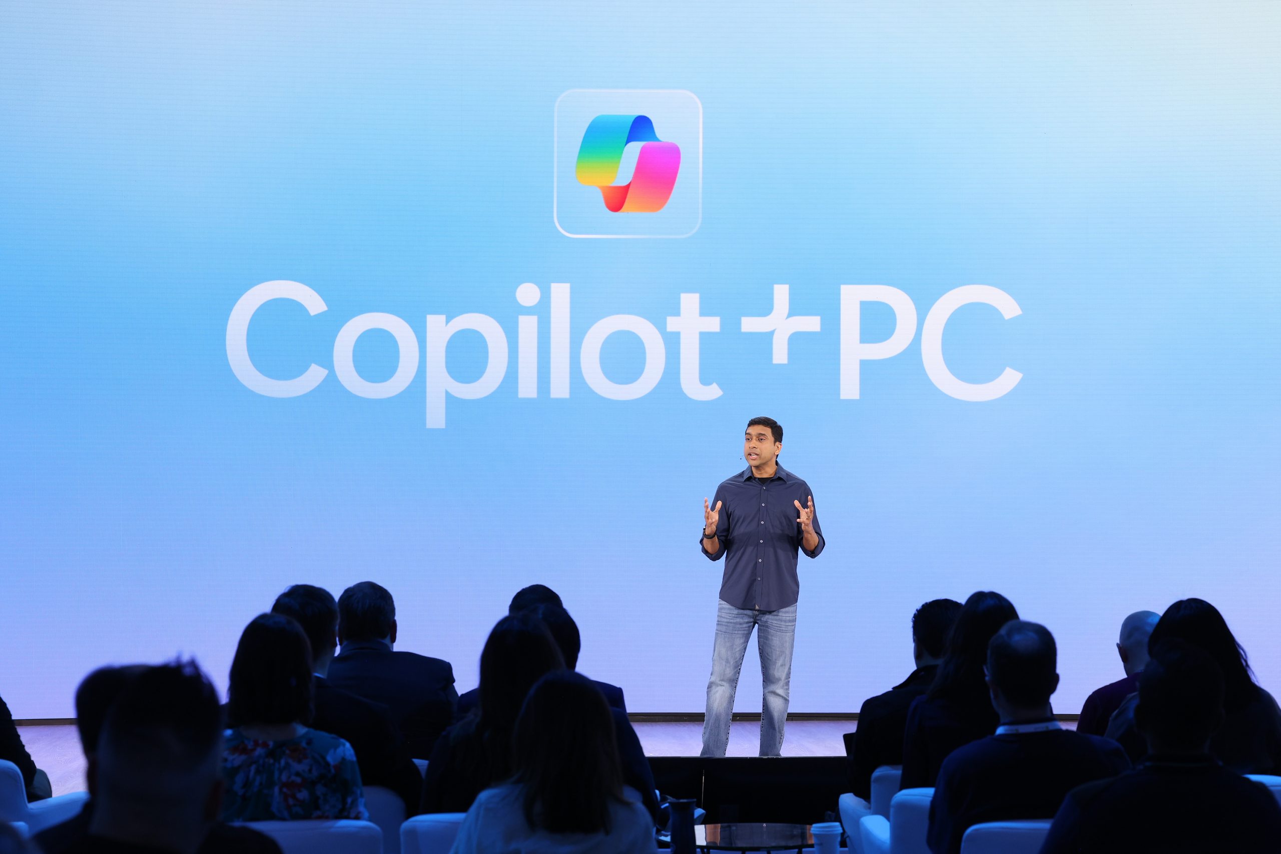 A man standing on stage in front of an audience, with the Copilot+ PC logo on the screen behind him