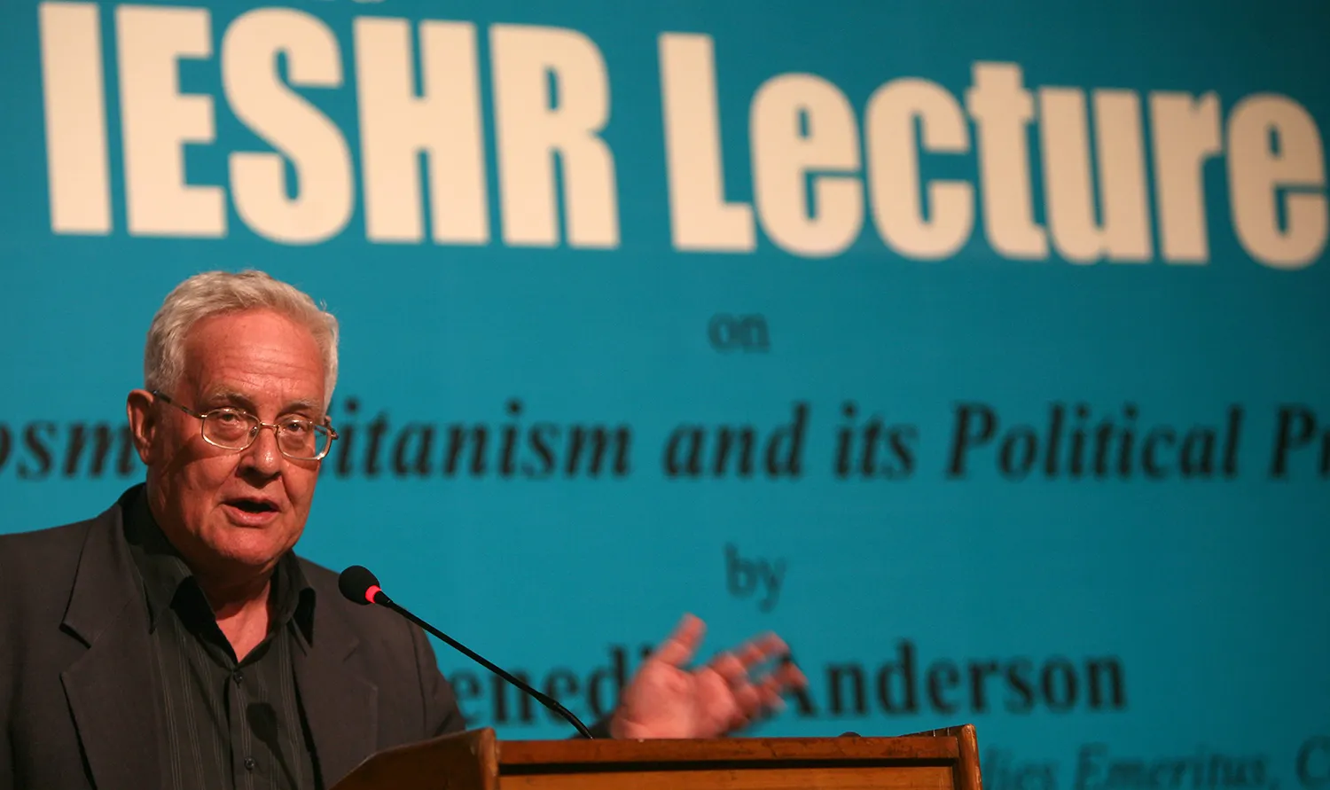 Intellectual and author Benedict Anderson stands behind a microphone and lecturn as he delivers a lecture.