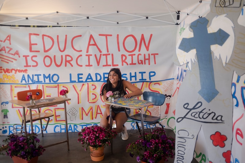 A young woman wearing shorts is seated outside at a student desk with a white banner hanging behind her that reads "Education is our civil right."