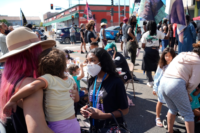 A young woman wearing a black T-shirt and a white face mask speaks with a mother with pink hair holding a baby.